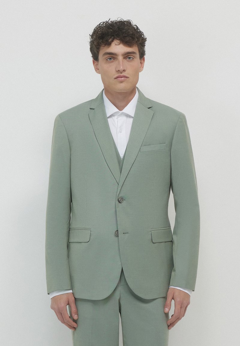Abito verde chiaro in tessuto strutturato, con chiusura a due bottoni, revers a punta e due tasche frontali. Indossato sopra una camicia bianca.