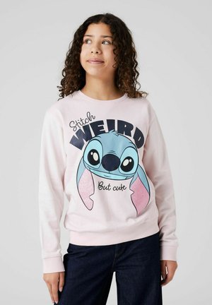 Ung person iført en lyserød sweatshirt med en stor blå tegneseriefigur og teksten "Stitch WEIRD But cute" på fronten.
