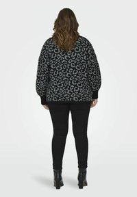 Femeie cu păr de lungime medie, îmbrăcată cu un pulover leopard negru și gri, pantaloni negri și cizmulițe cu toc negre, stând cu spatele.