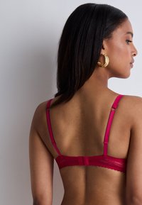 Soutien-gorge en dentelle rose avec des bretelles réglables et un bord festonné décoratif. La fermeture au dos est un design à deux crochets avec une finition lisse.