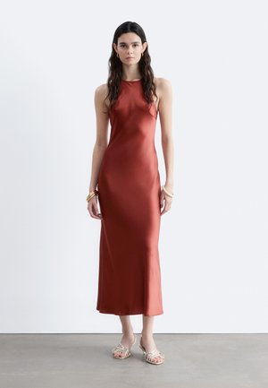 Mujer con cabello largo y oscuro, vestida con un vestido de satén sin mangas color óxido y sandalias doradas, de pie contra una pared blanca lisa.