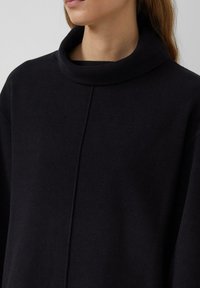 Czarny pulower z wysokim, miękkim kołnierzem oraz detalem w postaci przeszycia z przodu. Wykonany z teksturowanej tkaniny, prezentujący minimalistyczny design.