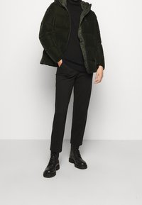 Tmavě zelená péřová bunda s kapucí, kterou doplňuje černý rolák a úzce střižené černé kalhoty. Na nohou má černé šněrovací boty s tlustou podrážkou.