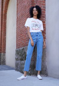 T-shirt graphique blanc avec un motif de crâne, jean bleu clair à jambes droites, et baskets blanches. Fond en briques et pierres texturées.