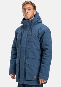 Mężczyzna w niebieskiej zimowej kurtce z kapturem, z dużymi kieszeniami z przodu i sznurkami, stojący na jednolitym tle.