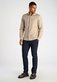 Licht beige overhemd met lange mouwen en een button-down kraag, gecombineerd met donkerblauwe broek en bruine leren schoenen. Klassieke pasvorm met subtiele textuur.