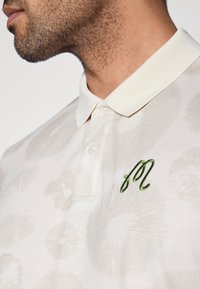 Balti polo marškinėliai su subtiliais gėlių raštais, apykakle ir žaliuoju siuvinėtu logotipu krūtinės srityje. Lygus tekstūra.