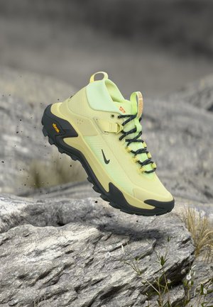 Scarpa da trekking verde chiaro e nera con robusta suola Vibram che levita sopra un terreno roccioso con piccole piante su uno sfondo grigio chiaro.