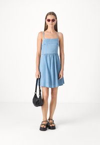 Robe en denim bleu clair avec de fines bretelles, design ajusté et évasé, et une taille froncée. Un sac bandoulière noir et des sandales plates complètent le look.