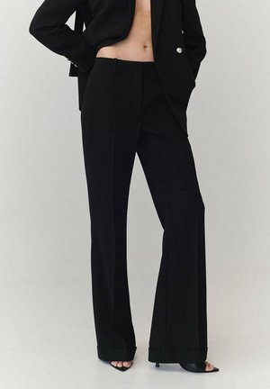 Mujer con pantalones negros de pierna ancha, blazer negro y tacones negros abiertos en los dedos, de pie con las manos en los bolsillos del blazer sobre un fondo liso.