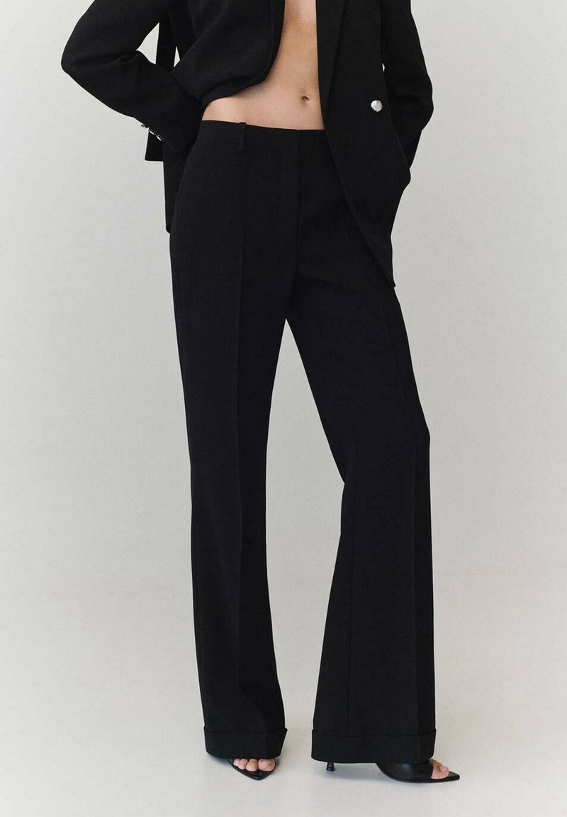 Femme portant un pantalon noir à jambes larges, un blazer noir et des talons noirs ouverts à l'avant, debout les mains dans les poches du blazer sur un fond uni.