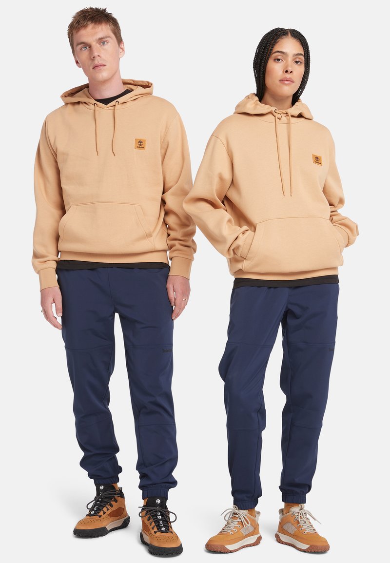Haut à capuche beige avec une poche avant et un logo, associé à un pantalon marine avec des poignets élastiques. Les deux portent des chaussures robustes de style extérieur.