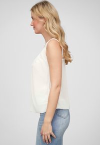 Haut sans manches blanc avec une texture lisse, présentant des bretelles délicates et un design ample et fluide. Associé à un jean ajusté bleu clair.