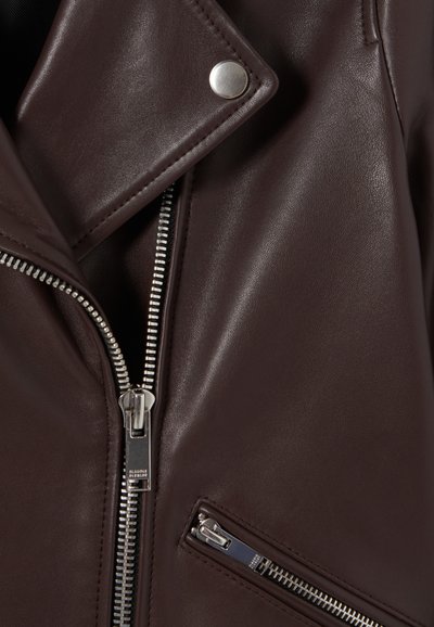 Veste en cuir marron style motard avec des revers en biais, des fermetures éclair argentées et des poches zippées. Texture lisse avec une finition légèrement brillante.