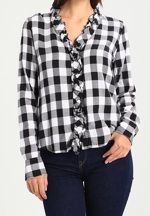 Frau trägt ein schwarz-weiß karierte Langarmhemd mit gerafften Vorderdetails, kombiniert mit dunkelblauen Jeans.