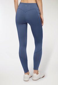 Niebieskie legginsy z wysokim stanem o gładkim, elastycznym wykończeniu, z wyprofilowanymi szwami i bocznymi kieszeniami, w zestawie z białymi butami sportowymi.