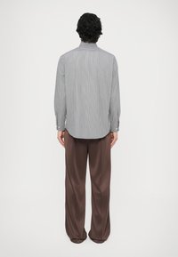 Srajca z dolgimi rokavi z belimi in svetlo sivimi črtami, s poudarjenim ukrivljenim robom, gumbi na manšetah ter sproščenim krojem, kombinirana z rjavimi širokimi hlačami.