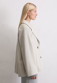 Femme blonde portant un manteau oversize beige clair à double boutonnage avec des manches de pull gris et un pantalon gris, faisant face à droite contre un mur blanc uni.
