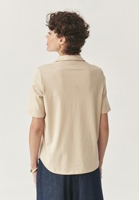 Camisa beige de manga corta hecha de tejido suave con cuello, que presenta un corte relajado y ligeras aberturas en los laterales del dobladillo.