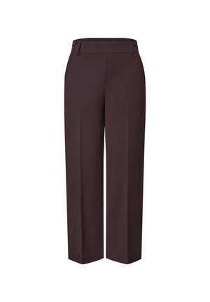 Femme portant un pantalon large marron foncé avec des poches latérales et des chaussures plates tissées marron, debout avec une main dans la poche.
