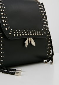 Musta nahkainen reppu, jossa on hopeiset niitit reunojen ympärillä. Repussa on läppä suljinta ja kaksi punottua hihnaa. Hopeiset metalliosat korostavat ilmettä.