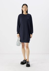 Abito felpa blu navy con maniche lunghe, polsini a costine e orlo; abbinato a scarpe con la piattaforma nere e una borsa a tracolla azzurro chiaro.