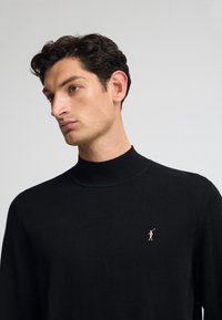 Svart stickad tröja med hög hals, med en liten logotyp i roséguld på vänster bröst. Mjuk texture och klassisk passform.