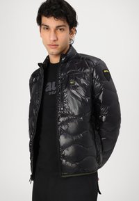 Giacca a vento nera con finitura lucida, dotata di chiusura lampo frontale, tasche laterali e un design trapuntato unico. Include una patch con logo.