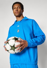 Μπλε αθλητική μπλούζα με μακριά μανίκια, γιακά και λογότυπο, κρατώντας μια ασπρόμαυρη μπάλα ποδοσφαίρου με αστέρι και πράσινες λεπτομέρειες.