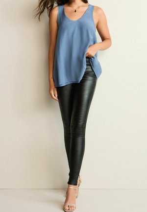 Femme portant un top bleu ample sans manches, des leggings en similicuir noir et des talons ouverts beige, debout contre un mur clair.
