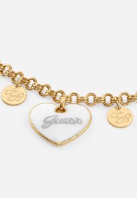 Gold-tone chain bracelet featuring a white heart charm with "Guess" in gray, and two small round pendants with engraved designs.
