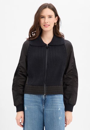 Frau trägt eine schwarze Jacke mit Reißverschluss, strukturiertem Vorderteil und aufgebauschten Ärmeln, kombiniert mit hellblauen, used Jeans.