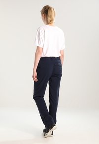 Person with blonde ponytail wearing a white short-sleeve shirt, navy blue pants, and black sneakers walking away on a light background.