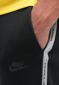Černé Nike Tech tepláky mají elegantní design s grafickým logem, bočními kapsami a bílým akcentním pruhem podél švů.