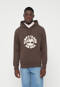 Jauns vīrietis, tērpies brūnā Timberland kapucē, virs gaiši rozā krekla un gaiši zilām džinsu biksēm, stāv uz vienkārša baltā fona.