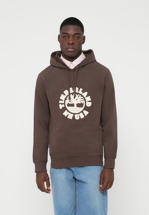 Junger Mann, der einen braunen Timberland-Hoodie über einem hellrosa Shirt und hellblauen Jeans trägt, steht vor einem schlichten weißen Hintergrund.