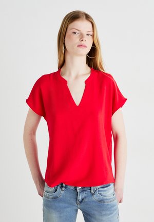 Blouse rouge à manches courtes avec un col en V, présentant une coupe décontractée et une texture douce. Associée à un jean bleu clair, créant un look décontracté.