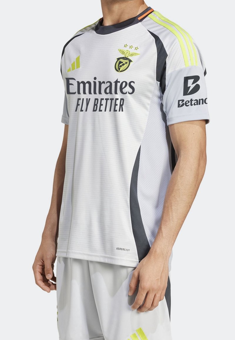 Camiseta de fútbol gris y negra con mangas cortas, que presenta un cuello redondo, acentos amarillos y gráficos del logo en el pecho y la manga.