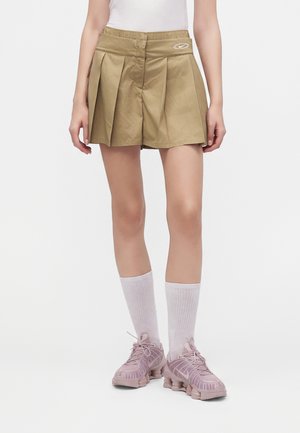 Personne portant un short plissé kaki, un haut blanc, des chaussettes blanches hautes et des chaussures de sport roses, debout sur un fond blanc.