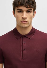 Burgundin värinen polo-paita, joka on valmistettu teksturoidusta kankaasta, jossa on kolmen napin halkio ja hillitty logotyyli edessä ja rinnassa.