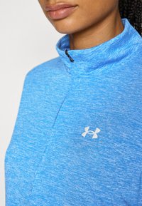 Blaues Sportoberteil mit einem Viertelreißverschlusskragen, aus einem strukturierten Stoff und mit einem weißen Logo auf der Brust. Nahaufnahme des Kragens.