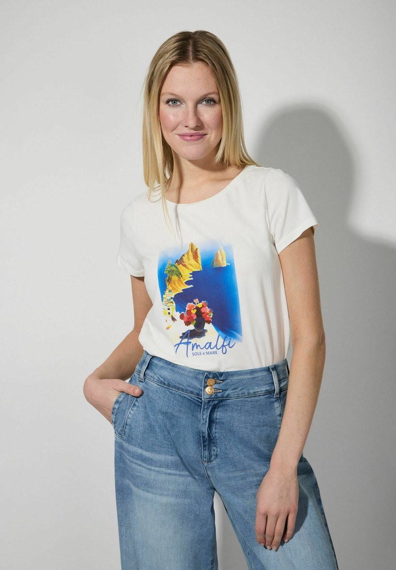 Weißes Baumwoll-T-Shirt mit einem bunten Amalfi-Design, das Blumen und Küstenlinie zeigt, kombiniert mit hellblauen, weiten Jeans mit zwei Knöpfen.