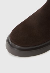 Bruine suède enkelboot met een ronde neus en een contrasterende zwarte rubberen zool, die de zachte structuur en het eenvoudige ontwerp benadrukt.