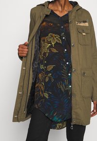 Parka verde oliva con capucha, con herrajes plateados y un bolsillo frontal. Debajo, una camisa de estampado floral oscuro en tonos azules y verdes.