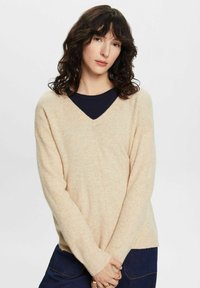 V-hals trui in lichtbeige, gemaakt van zacht materiaal. Lange mouwen, iets oversized pasvorm, geribbelde manchetten en onderrand, gladde textuur.