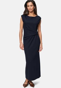 Navyblaues, ärmelloses Maxikleid mit einem geknoteten Detail an der Taille, aus einem glatten Stoff gefertigt. Braune Schuhe ergänzen das Outfit.