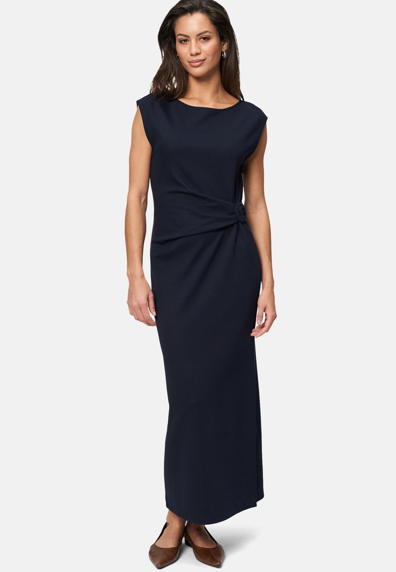 Navyblaues, ärmelloses Maxikleid mit einem geknoteten Detail an der Taille, aus einem glatten Stoff gefertigt. Braune Schuhe ergänzen das Outfit.