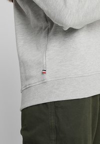 Jasnoszara bawełniana bluza z prążkowanym dołem, z małą czerwoną, białą i granatową metką z logo po bokach. W połączeniu z ciemnozielonymi spodniami.