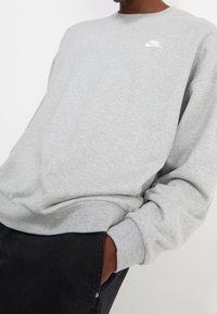 Grå sweatshirt i mjukt tyg, med rund hals, långa ärmar och vit Nike-logotyp på bröstet. Bärs med svarta byxor.