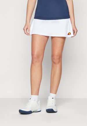 Witte tennisrok met een gestructureerd ontwerp, gecombineerd met witte sportschoenen met marineblauwe accenten en logo. Donkere blauwe top zichtbaar erboven.